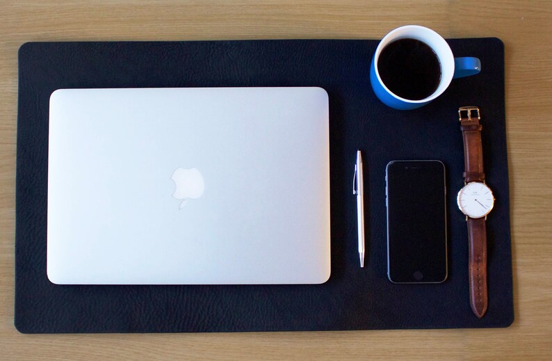 Puede incluir: Una alfombrilla de escritorio de cuero negro con un port&aacute;til, un tel&eacute;fono inteligente, un bol&iacute;grafo, una taza de caf&eacute; azul y un reloj de pulsera con una correa de cuero marr&oacute;n.