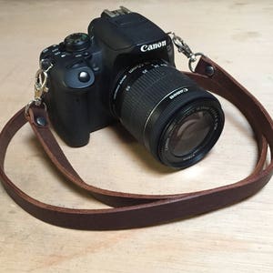 Könnte beinhalten: Ein braunes Lederkameraband mit silbernen Verschlüssen, das an einer schwarzen Canon DSLR-Kamera mit einem Objektiv befestigt ist.