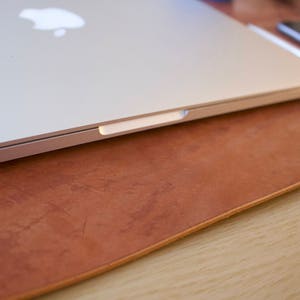 Puede incluir: Un ordenador port&aacute;til plateado cerrado que descansa sobre una alfombrilla de escritorio de cuero marr&oacute;n. El port&aacute;til tiene un logotipo de Apple en la tapa.