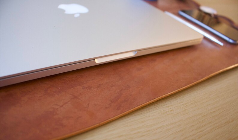 Puede incluir: Un ordenador port&aacute;til plateado cerrado que descansa sobre una alfombrilla de escritorio de cuero marr&oacute;n. El port&aacute;til tiene un logotipo de Apple en la tapa.