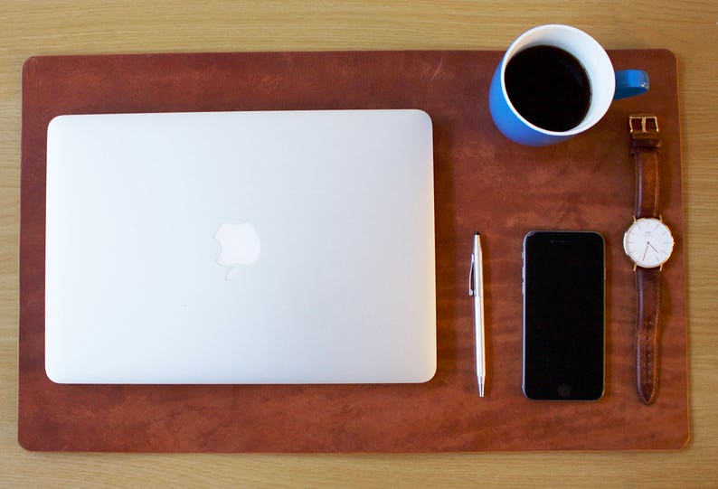 Puede incluir: Una alfombrilla de escritorio de cuero marr&oacute;n con un port&aacute;til, un tel&eacute;fono inteligente, un bol&iacute;grafo, una taza de caf&eacute; azul y un reloj de pulsera con una esfera blanca.