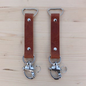 Puede incluir: Dos correas de cuero marrón con bucles y clips de metal plateado. Las correas cuelgan de una superficie de madera.