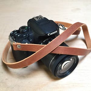 May include: A brown leather camera strap with a Canon camera attached. The camera is black with a silver lens.