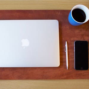 Puede incluir: Una alfombrilla de escritorio de cuero marr&oacute;n con un port&aacute;til plateado, una taza de caf&eacute; azul, un bol&iacute;grafo plateado, un tel&eacute;fono inteligente negro y un reloj de cuero marr&oacute;n con una esfera blanca.