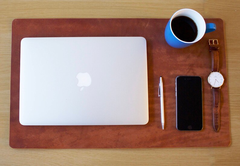 Puede incluir: Una alfombrilla de escritorio de cuero marr&oacute;n con un port&aacute;til plateado, una taza de caf&eacute; azul, un bol&iacute;grafo plateado, un tel&eacute;fono inteligente negro y un reloj de cuero marr&oacute;n con una esfera blanca.