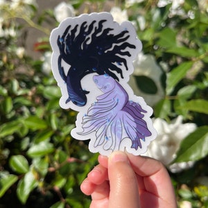 May include: A sticker featuring two betta fish, one black and one purple, with flowing fins. The fish are facing each other, creating a heart shape.
