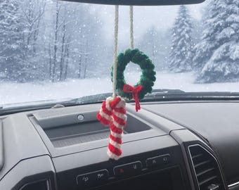 May include: A festive car decoration comprising a green crocheted wreath with a red bow and a red and white striped candy cane. The charm hangs from a white string, with a snowy forest visible through the windscreen.