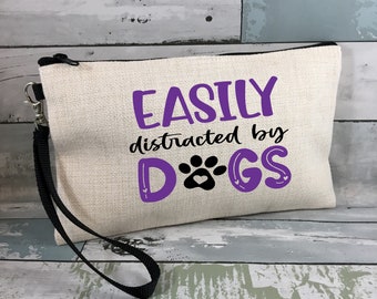 May include: A white canvas zippered pouch with a black strap. The pouch has the text "EASILY distracted by DOGS" in purple and black lettering with a paw print.