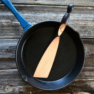May include: A black cast iron skillet with a blue handle and a wooden spatula with a black handle. The spatula has a carved design on the handle.