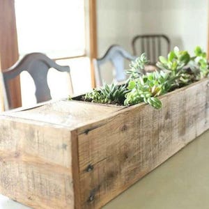 May include: A rectangular wooden planter box filled with green succulents. The box is made of weathered wood, showcasing natural grain and knots. The planter is a decorative item for indoor or outdoor use.