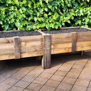 May include: A wooden raised garden bed with two sections, filled with soil. The bed is made of reclaimed wood and has a rustic look. It is sitting on a patio made of brown tiles.