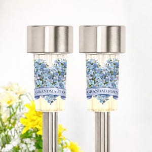 May include: Two solar garden lights with stainless steel tops. The lights feature a floral design with blue flowers and the text "GRANDMA FLO" and "GRANDAD JOHN" on a banner. The lights are set against a white background.