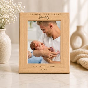 May include: A light oak picture frame with the text "THE MOMENT YOU BECAME MY Daddy" at the top. The frame contains a photo of a person holding a newborn baby. The bottom of the frame has the date, time, and name.