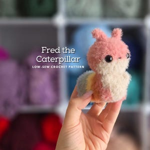 May include: A pink and white crocheted caterpillar toy named Fred. The toy has a fuzzy texture and black eyes. The text "Fred the Caterpillar" and "Low-Sew Crochet Pattern" is visible in the image.