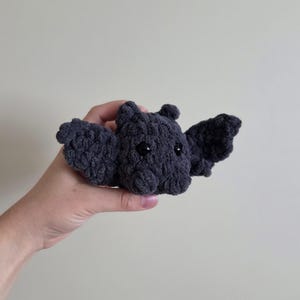 May include: A dark gray crocheted bat with black eyes and outstretched wings. The handmade plush toy is held in a hand against a neutral background. The bat's wings are spread, and its body is textured.