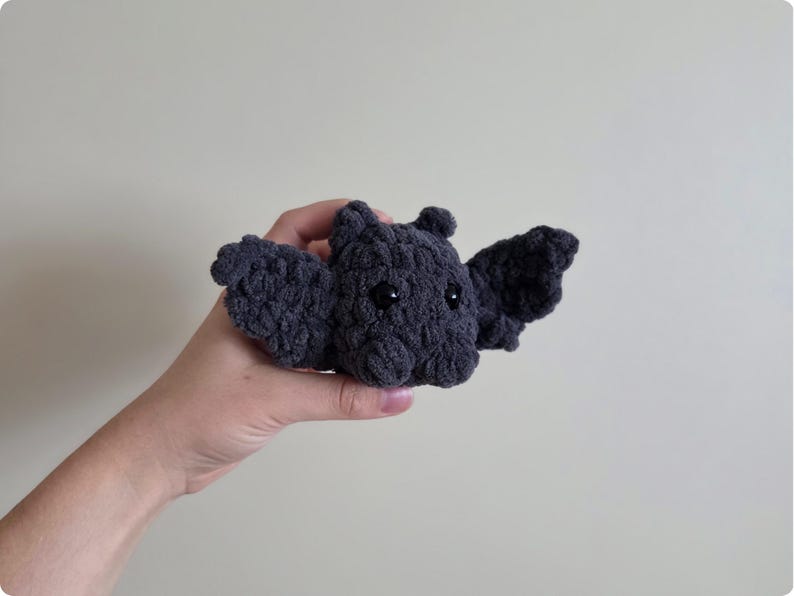 May include: A dark gray crocheted bat with black eyes and outstretched wings. The handmade plush toy is held in a hand against a neutral background. The bat's wings are spread, and its body is textured.