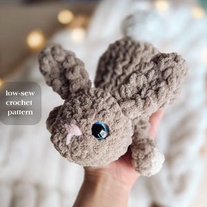 May include: A handmade, plush bunny in shades of brown, with large blue eyes and a pink nose. The bunny has long ears and is made from a soft, textured material. The text "low-sew crochet pattern" is visible.