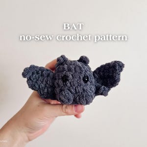 Pode incluir: Um morcego de crochê cinza escuro feito à mão, com olhos pretos e asas abertas. O texto "BAT no-sew crochet pattern" é exibido acima do morcego. O morcego é segurado em uma mão.