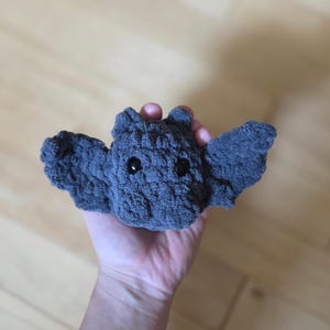 May include: A handmade gray crochet bat with black safety eyes and outstretched wings. This plush toy is a cute and cuddly creature, perfect for Halloween or as a unique gift. The bat is made of soft, textured yarn.