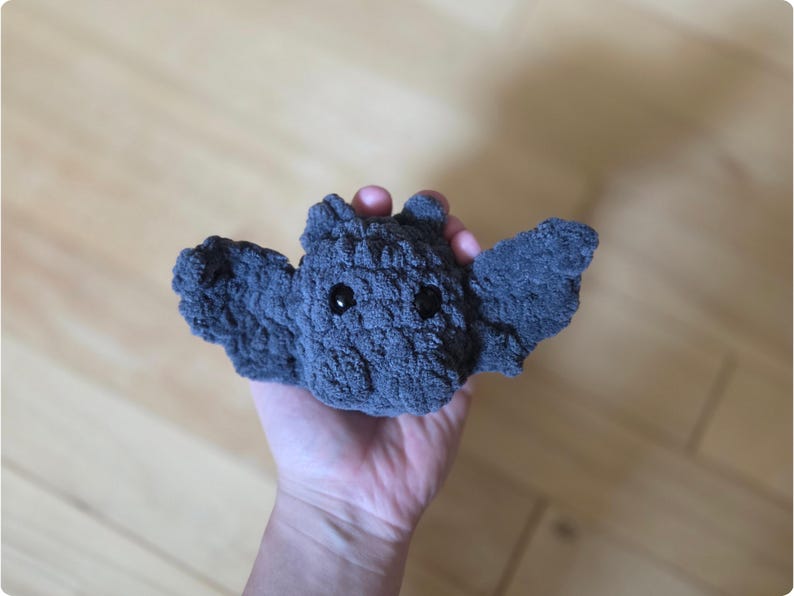 May include: A handmade gray crochet bat with black safety eyes and outstretched wings. This plush toy is a cute and cuddly creature, perfect for Halloween or as a unique gift. The bat is made of soft, textured yarn.