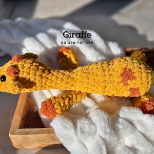 May include: A crocheted giraffe toy with a yellow and orange color scheme. The giraffe is lying on a white fluffy blanket. The text "Giraffe NO-SEW PATTERN" is visible above the giraffe.