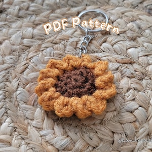 May include: A crocheted sunflower keychain with a brown center and yellow petals. The keychain is attached to a silver key ring. The text "PDF Pattern" is written in white on a black background above the keychain.