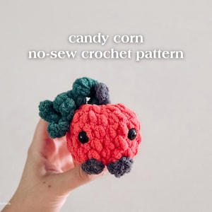 May include: A handmade, red crocheted candy corn with black eyes and green and gray accents. The text "candy corn no-sew crochet pattern" is displayed above the candy corn. The candy corn is held in a hand.