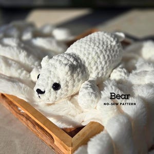 May include: A white crocheted bear toy on a white fluffy blanket. The bear has black eyes and a black nose. The blanket is in a wooden tray. The text "Bear No-Sew Pattern" is on the blanket.