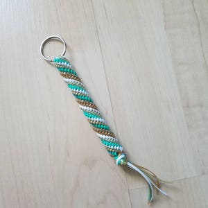 May include: A handmade keychain with a braided design. The keychain features a silver metal ring and a braided cord in shades of teal, white, and gold. The end of the cord has a small knot with several strands of white, teal, and gold cord.