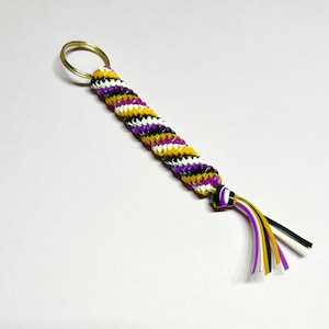 Pode incluir: Um chaveiro trançado roxo, branco e amarelo com um aro de chave dourado. A trança é torcida e tem detalhes pretos.