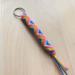 May include: A colorful key chain with a braided design in blue, yellow, pink, and white. The key chain has a silver key ring at the top and a tassel at the bottom.