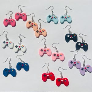 Puede incluir: Un conjunto de 10 pares de pendientes de mando de videojuegos de colores. Los pendientes están hechos de arcilla polimérica y presentan una variedad de colores, incluyendo rosa, azul, blanco, negro, rojo y morado.