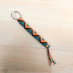 May include: A colorful braided keychain with a silver key ring. The keychain is made with black, orange, and turquoise cords and has a tassel at the end.