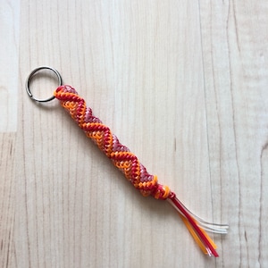 May include: A handmade keychain with a spiral design in shades of red, orange, and pink. The keychain has a silver metal ring and clear plastic strands with red and orange accents at the end. The keychain is laying on a light wood surface.