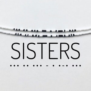 Könnte beinhalten: Zwei weiße und schwarze Perlenarmbänder mit dem Wort "SISTERS" darunter gedruckt. Das Wort "SISTERS" ist in Großbuchstaben und in einer schwarzen serifenlosen Schrift.