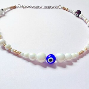 Puede incluir: Un collar de perlas blancas con cuentas doradas y dijes de ojo turco azul.