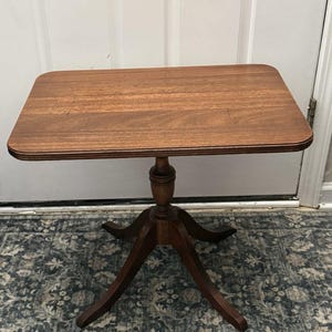 Puede incluir: Una mesa auxiliar rectangular de madera con un acabado liso en color marrón medio. El tablero tiene esquinas redondeadas y un borde escalonado. Se apoya en una columna central que termina en una base de cuatro patas curvadas.
