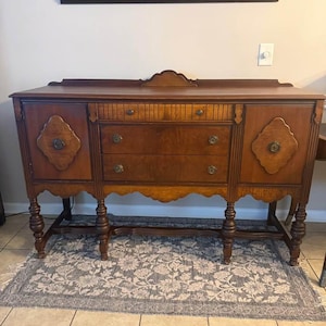 May include: A dark wood antique buffet with three drawers and two side cabinets. The buffet has ornate details, including decorative diamond-shaped panels and brass hardware. The piece is supported by turned legs and sits on a floral patterned rug.