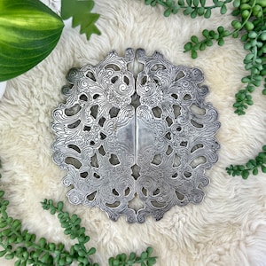 May include: A silver metal trivet with a decorative floral design. The trivet is made of two pieces that are joined together in the center. The trivet is sitting on a white fluffy surface.