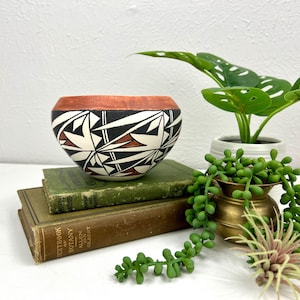May include: A handcrafted ceramic bowl with a geometric design. The bowl features a white base with black, red, and brown patterns. The rim is painted a terracotta color. The bowl is displayed on top of two vintage green books.