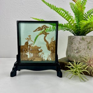 May include: A framed, three-dimensional artwork depicting a detailed Asian landscape scene. The artwork features a pagoda, trees, and mountains, all crafted from a light brown material. The frame is black, and the artwork is displayed on a black stand.