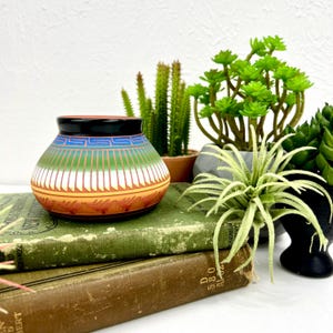 May include: A small, hand-painted ceramic pot with a black rim and a colorful design. The pot features bands of blue, green, orange, and white, with geometric patterns. It is displayed on top of vintage books, with green plants in the background.