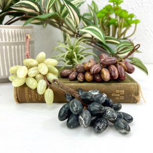 May include: Three bunches of grapes made of stone. The grapes are green, brown, and black. The grapes are sitting on an old book.