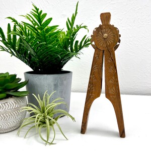 May include: A wooden nutcracker with intricate carved floral designs. The nutcracker is brown and has a circular design at the top. The legs of the nutcracker are open. The background includes green plants in gray and patterned pots.