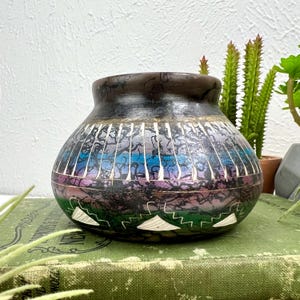 May include: A handcrafted, decorative ceramic pot with a dark brown rim and a colorful, patterned body. The design features shades of blue, purple, and green with white accents. The pot has a rounded shape and is likely used for display or storage.