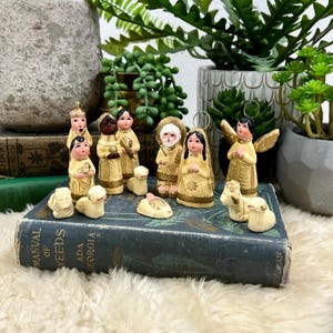 May include: A collection of hand-painted Christmas nativity figurines. The figures are in cream and gold tones, depicting Mary, Joseph, the baby Jesus, angels, and shepherds. The figurines are arranged on a vintage book.