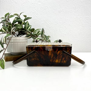 May include: A brown and clear plastic box purse with a tortoise shell pattern and two black handles.