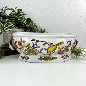 May include: White ceramic planter with a rectangular shape and floral and bird design. The planter features a yellow bird, red, yellow, and green flowers, and brown handles. The planter is a decorative piece for indoor plants.