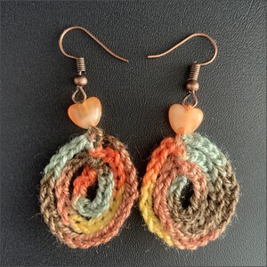 May include: A pair of handmade crochet earrings. Each earring features a spiral design in shades of brown, orange, yellow, and green. A small, heart-shaped bead in a peach color hangs below the copper ear wire.