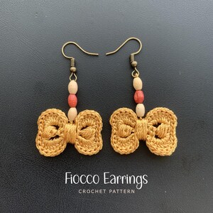 May include: A pair of handmade, gold-colored crochet bow earrings. Each earring features a bow shape with a textured design, hanging from a metal hook. The earrings are adorned with wooden beads in cream and red. The text "Fiocco Earrings CROCHET PATTERN" is at the bottom.